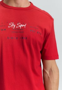 Rotes T-Shirt mit kurzen Ärmeln aus Baumwolle. Mit weißem Markenaufdruck und blauen Druckdetails. Klassischer Rundhalsausschnitt und lockere Passform.