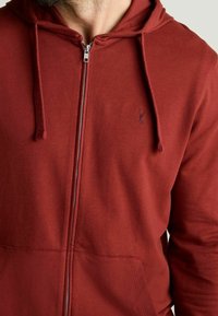 Sudadera roja con cremallera, hecha de un tejido suave, que cuenta con un bolsillo frontal, capucha con cordón y un pequeño logo en el pecho.