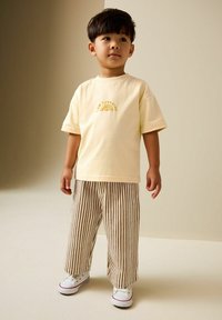 Junge steht und trägt ein cremefarbenes T-Shirt mit dem Text "hello sunshine", braun-weiß gestreifte Hosen und weiße Turnschuhe vor einem neutralen Hintergrund.