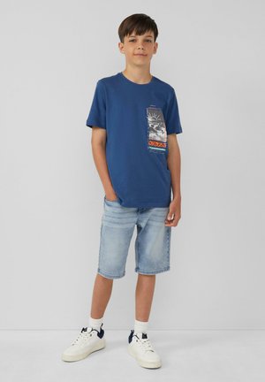 Blauwe katoenen t-shirt met een grafische zakprint, gecombineerd met lichte denim shorts. Witte sneakers met marineaccenten maken de outfit compleet.