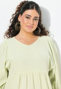 Ljusgrön, textured blus med V-ringning, puffärmar och rynkad midja. Guldörhängen i form av ringar ger detalj.