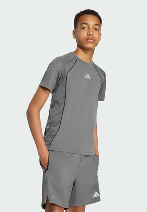 Jeune homme portant un t-shirt de sport gris et un short avec des logos Adidas, debout les mains dans les poches sur un fond uni.