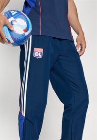 Calças de desporto azul-marinho com acentos vermelhos, apresentando riscas laterais brancas e um logótipo do Olympique Lyonnais. Mão segurando uma bola de futebol azul com padrão.