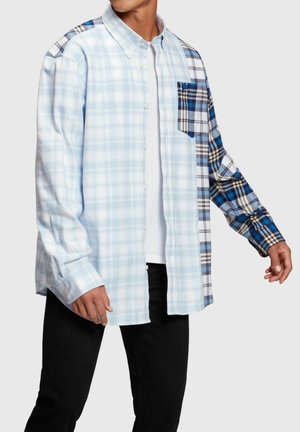 Camicia a quadri blu con bottoni, caratterizzata da una combinazione di quadretti blu chiaro e blu scuro. Un lato presenta una tasca a quadri contrastante. Materiale misto cotone.