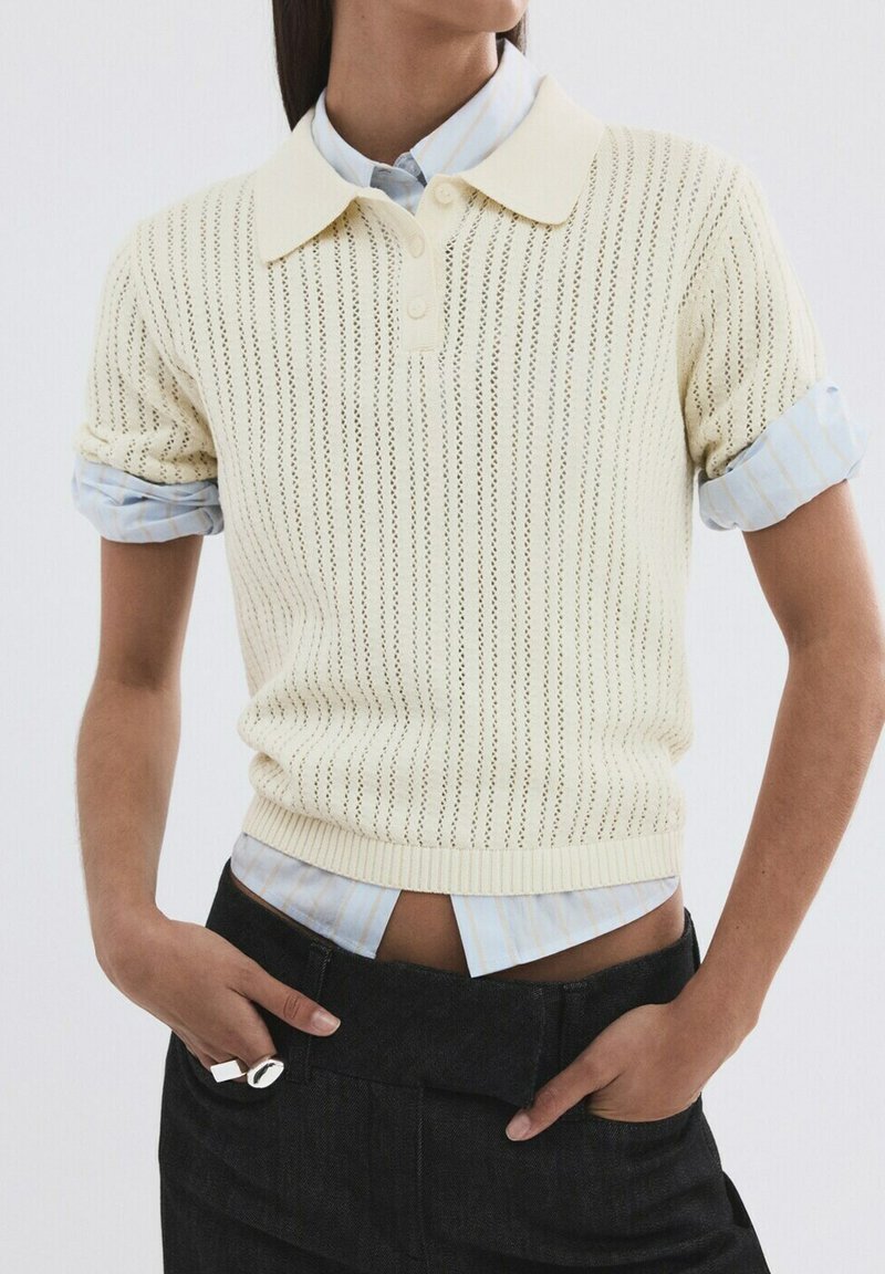 Person wearing a cream knit polo sweater over a light blue striped button-up shirt with sleeves rolled, paired with black high-waist pants.
