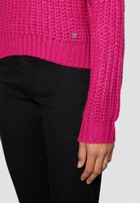 Fuksianvärinen neulottu pusero, jossa on teksturoitu kuvio ja resorireunukset, yhdistetty mustiin istuviin housuihin. Pieni metallinen logo sivussa.