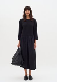 Schwarzes langärmliges Kleid mit eng anliegendem Oberteil und gerafftem Rock. Getragen mit schwarzen Slippers; hält eine große schwarze Ledertasche.