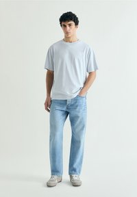 Joven de pie con una mano en el bolsillo, vestido con una camiseta holgada de color gris claro, vaqueros azul claro y zapatillas grises sobre un fondo liso.