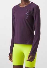 Fioletowa długosleeve'owa koszulka sportowa z okrągłym dekoltem i nadrukowanym logo. W połączeniu z jaskrawo neonowymi żółtymi dopasowanymi szortami. Gładka tekstura, elastyczny materiał.