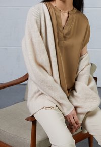 Beige gestrickte Strickjacke über einer braunen Bluse mit Knopfleiste, kombiniert mit cremefarbenen Hosen. Weiche Texturen und lockere Passform, auf einem Stuhl sitzend.