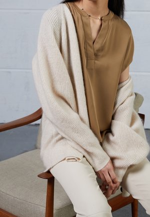 Un cardigan beige tricoté sur une blouse marron avec une patte de boutonnage, associé à un pantalon crème. Textures douces et coupe décontractée, assise sur une chaise.
