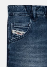 Σκούρες μπλε τζιν παντελόνες με μια μικρή μπροστινή τσέπη που φέρει ετικέτα με λογότυπο και ορατές ραφές. Ο ιστός έχει μια λεία, ελαφρώς φθαρμένη υφή.