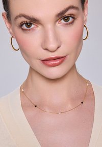 Złoty naszyjnik z łańcuchem z małymi czarnymi i białymi koralikami, noszony z złotymi kolczykami w kształcie kół z perłowymi akcentami. Gładka karnacja i neutralna bluzka.