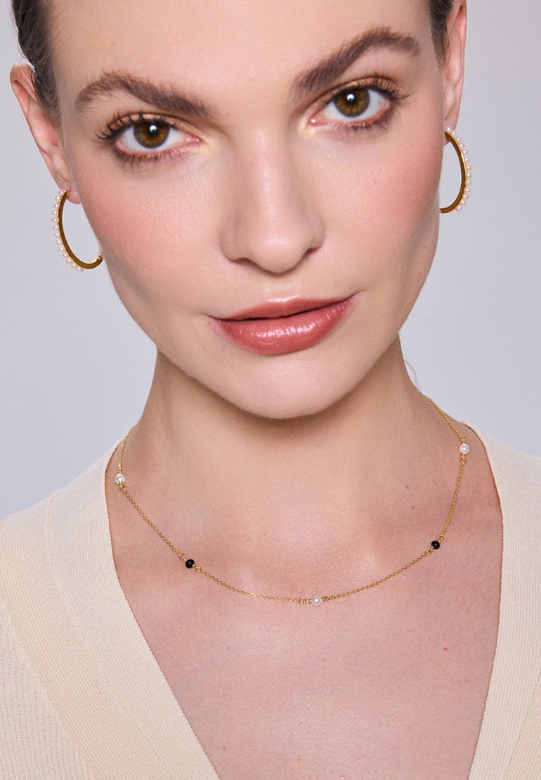 Złoty naszyjnik z łańcuchem z małymi czarnymi i białymi koralikami, noszony z złotymi kolczykami w kształcie kół z perłowymi akcentami. Gładka karnacja i neutralna bluzka.