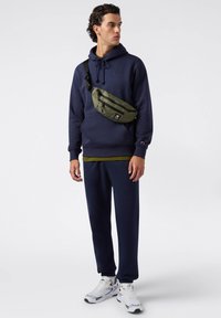 Felpa blu scura, marsupio verde oliva, pantaloni jogger blu e sneakers bianche. Il marsupio è dotato di chiusure lampo e tracolla regolabile.