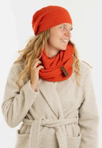 Oranje gebreide muts met een textuurpatroon, gecombineerd met een bijpassende oneindige sjaal. Het model draagt een lichtgekleurde, fluffy jas.