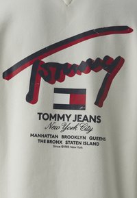 Crème kleurige sweatshirt met een groot rood en zwart 'Tommy' logo boven een rechthoekig logo in blauw, wit en rood. Tekst eronder geeft locaties en datum aan.