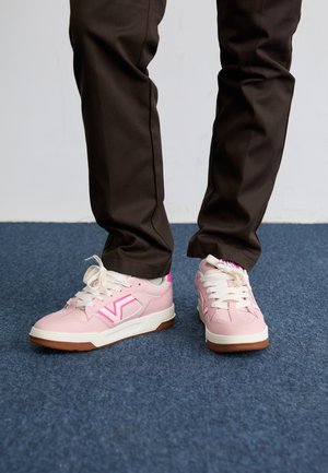 Zapatillas rosas con una textura suave, suela blanca y suela de goma marrón. El diseño presenta un logo prominente en rosa en el lado.