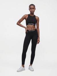 Γυναίκα που φοράει μαύρο crop top και κολάν με το κείμενο "Palm Angels", σε συνδυασμό με λευκά αθλητικά παπούτσια, σε λευκό, απλό φόντο.