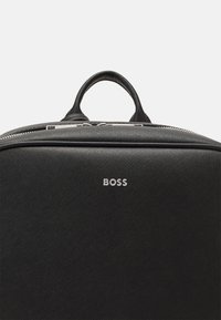 Black leather backpack with a textured surface, featuring a top handle and a silver logo. Equipped with a zipper closure and sleek design.