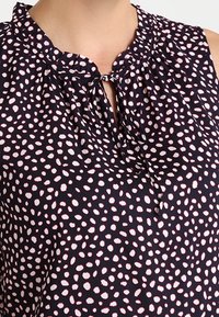 Detailní záběr na bezrukávovou tmavě modrou blůzu s bílými nepravidelnými tečkami a krčním výstřihem ve tvaru klíčové dírky, který je svázán tenkou látkovou mašlí.