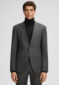 Mann in einem grauen Nadelstreifenanzug mit einem schwarzen Rollkragenpullover, der vor einem schlichten hellgrauen Hintergrund steht.