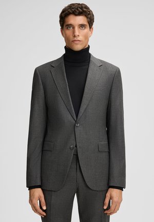 Mann in einem grauen Nadelstreifenanzug mit einem schwarzen Rollkragenpullover, der vor einem schlichten hellgrauen Hintergrund steht.