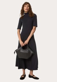 Schwarzes, mittellanges Kleid mit kurzen Ärmeln und hohem Kragen. Das Model hält eine strukturierte, schwarze Handtasche mit verzierten Griffen. Schwarze flache Schuhe.