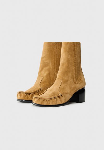 Bottines en suede marron clair avec un bout arrondi, des panneaux latéraux élastiques et un large talon noir. Présente des détails cousus le long des bords.