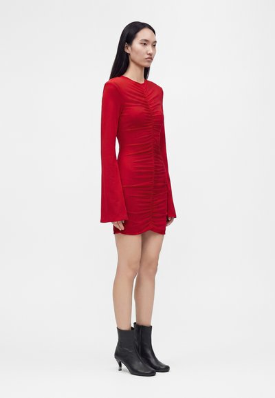 Femme aux longs cheveux foncés porte une robe mini rouge froncée avec des manches longues évasées et des bottines noires à talons, debout devant un fond uni.