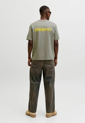 Mand iført solbriller, grå Jack & Jones t-shirt med gult logo, mørke løse jeans og beige sko, stående med ryggen til på hvid baggrund.