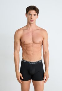 Boxeri scurți negri din bumbac, cu o potrivire strânsă, având un bărbierit elastic cu marcă de culoare albă. Modelul are o constituție atletică și stă cu fața spre cameră.
