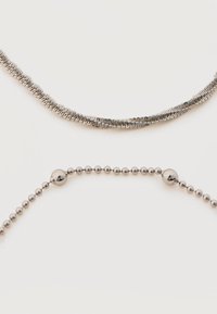 Δύο ασημένια μεταλλικά κολιέ, το ένα με επίπεδο ζιγκ-ζαγκ μοτίβο και το άλλο με μικρές στρογγυλές χάντρες και μεγαλύτερες διακοσμητικές χάντρες.