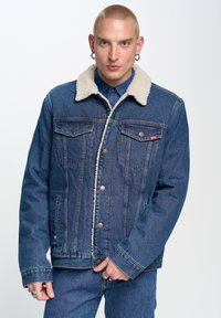 Denimjakkk with shearling kraega, nööpide sulgemine ja kaks rinna taskut. Pehme tekstuur, tume sinine värv ja nähtavad õmblusdetailid.