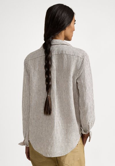 Sieviete ar garām pinētām tumšām matiem, valkājot brūnu un baltu vertikāli svītrainu garroku kreklu un bēša krāsas bikses, redzama no muguras.