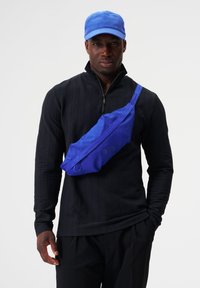 Blaues sling Bag aus Stoff mit glatter Textur, ausgestattet mit einem Reißverschluss und kreisförmigen Akzenten. Wird über einem schwarzen Langarmshirt getragen.