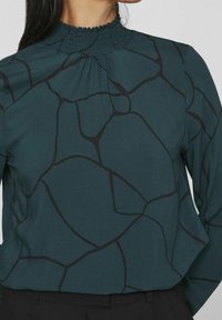 Blusa verde acqua con colletto alto arricciato, caratterizzata da un motivo astratto nero. Il tessuto ha una superficie liscia, con maniche lunghe e un taglio morbido.