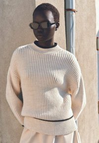 Gestrickter cremefarbener Pullover mit geripptem Muster, Rundhalsausschnitt und schwarzem Akzent am unteren Rand. Modell trägt reflektierende Sonnenbrille.
