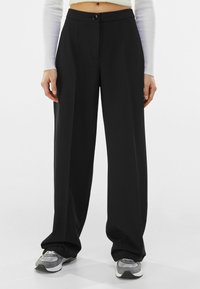 Černé kalhoty širokého střihu vyrobené z hladké tkaniny, s předním zapínáním na knoflík, poutky na opasek a bočními kapsami, doplněné šedými teniskami.