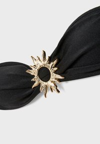 Czarna tkaninowa opaska do włosów przekręcona przez złoty metalowy pierścień w kształcie słońca z teksturowanymi, przypominającymi płomienie promieniami.
