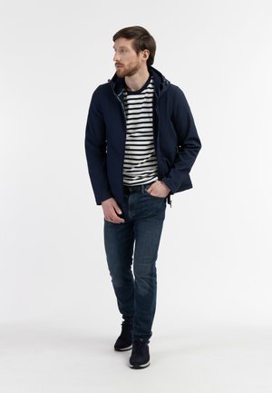 Mann trägt marineblaue Jacke, weiß-blau gestreiftes Hemd, blaue Jeans und schwarze Sneakers, geht und schaut zur Seite vor weißem Hintergrund.