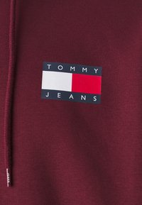 Felpa bordeaux con logo rettangolare caratterizzato da blocchi blu navy, bianchi e rossi. Il materiale ha una texture liscia con dettagli a coste.