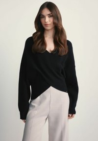 Czarny sweter dzianinowy o kroju z zawinięciem i dekolcie w kształcie litery V, w zestawieniu z jasnymi, dopasowanymi spodniami. Gładka tekstura, luźny krój.