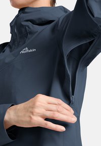 Giacca impermeabile blu navy con una tasca laterale con zip e cappuccio regolabile, caratterizzata da una texture liscia e un logo riflettente.