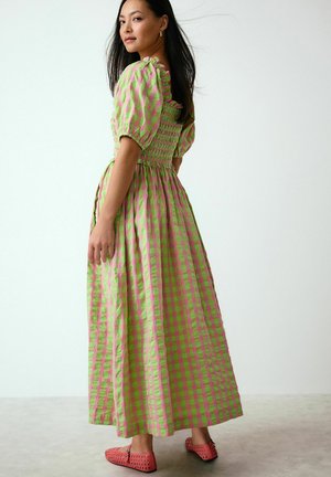 Woman wearing green and pink checkered puff-sleeve maxi dress and coral perforated slip-on shoes, standing with head turned over shoulder.