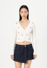 Cardigan bianco a coste con motivi a cuore rossi, caratterizzato da un profondo scollo a V e chiusura con bottoni. Abbinato a una mini gonna in denim scuro.