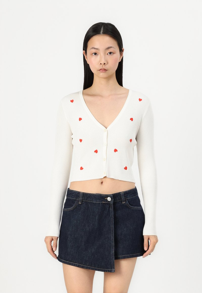Cardigan bianco a coste con motivi a cuore rossi, caratterizzato da un profondo scollo a V e chiusura con bottoni. Abbinato a una mini gonna in denim scuro.