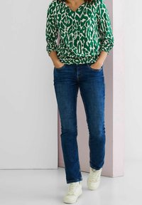 Donna che indossa una maglietta verde e bianca con stampa animale, jeans blu e sneakers bianche, in piedi con le mani nelle tasche su uno sfondo neutro.