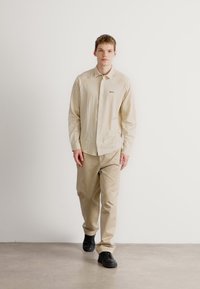 Lichtbeige gestreepte overhemd met lange mouwen en knoopsluiting, gecombineerd met lichtkhaki broek en zwarte schoenen, staande tegen een witte achtergrond.