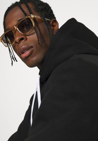 Black hooded sweatshirt with white drawstrings, paired with oversized light-brown sunglasses; features braided hair and neutral background.
